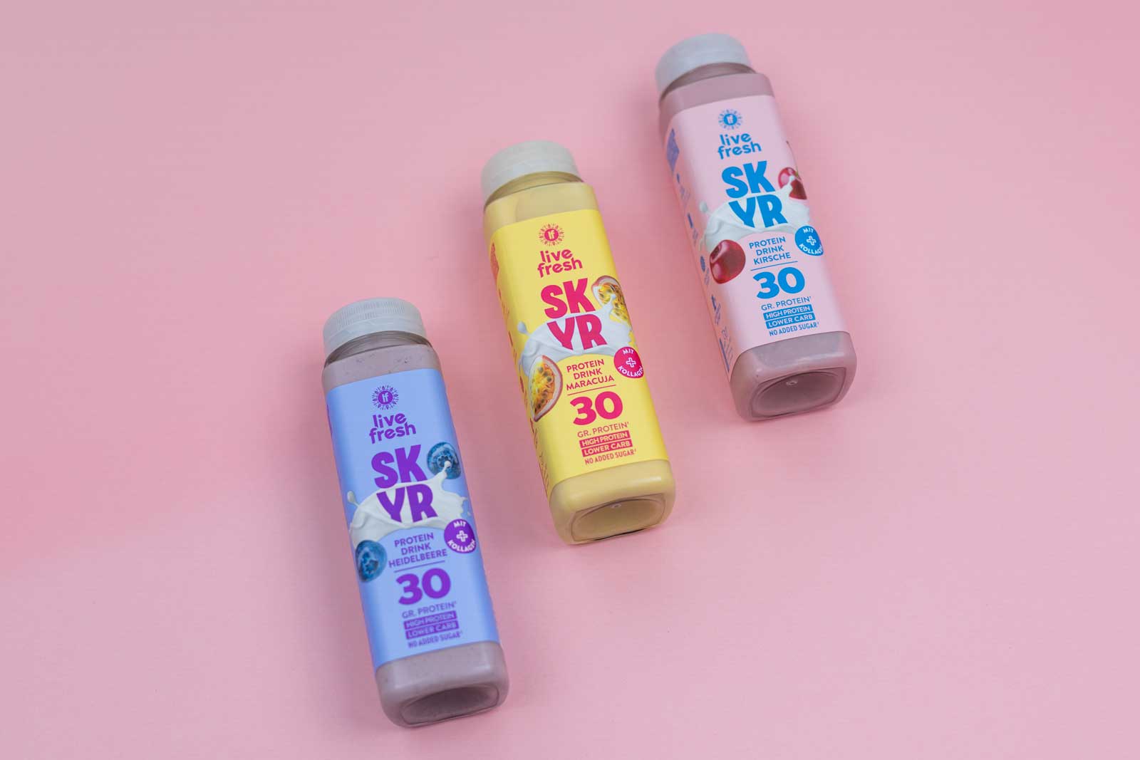 Drei Flaschen LiveFresh Skyr Protein Drink auf rosa Hintergrund, sortiert in den Sorten Heidelbeere (lila Flasche), Maracuja (gelbe Flasche) und Kirsche (rosa Flasche). Jede Flasche enthält 30 g Protein, ist zuckerfrei, low carb und mit Kollagen angereichert.