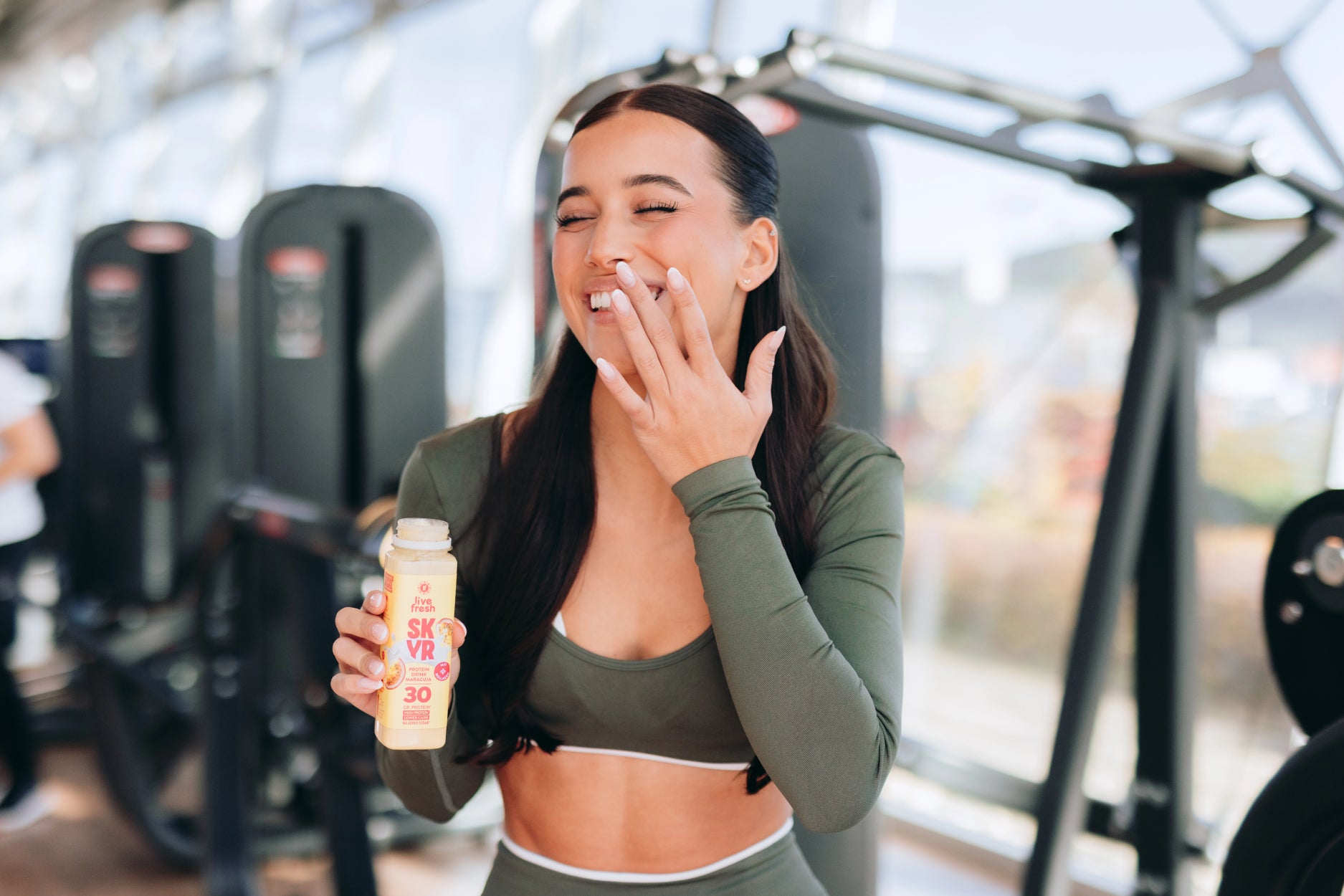  Lachende Frau im Fitnessstudio hält eine Flasche Proteinshake in der Hand und genießt einen Schluck nach dem Training.