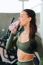 Frau in grünem Sportset trinkt einen rosa Proteinshake im Fitnessstudio neben Trainingsgeräten.