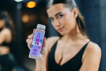 Frau in schwarzer Sportkleidung präsentiert eine Flasche LiveFresh SKYR Protein Drink Heidelbeere mit 30 Gramm Protein im Fitnessstudio.