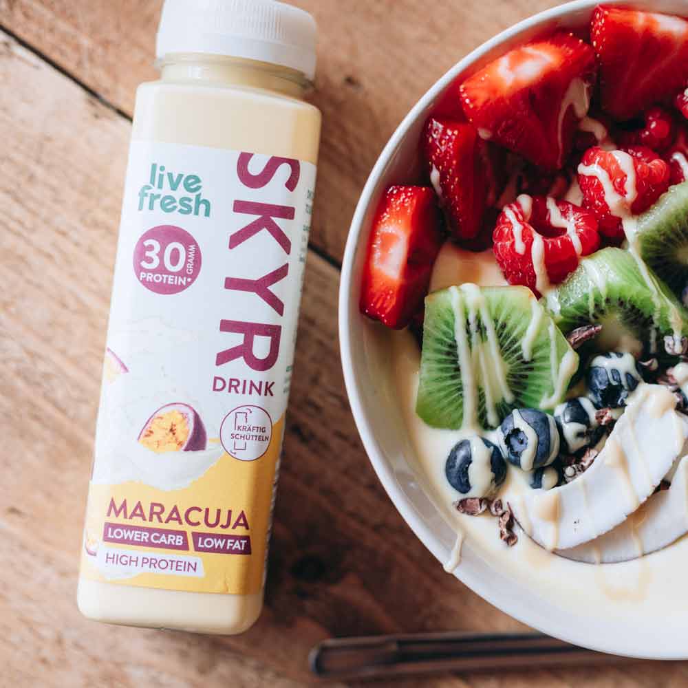 Eine Bowl, garniert mit Erdbeeren, Himbeeren, Kiwi, Blaubeeren und Kakaonibs. Daneben steht eine Flasche „Live Fresh Skyr Drink Maracuja“ mit 30g Protein. Die Skyr-Flasche wirbt mit niedrigem Kohlenhydratgehalt, fettarm und proteinreich, und betont eine gesunde und ausgewogene Ernährungswahl.