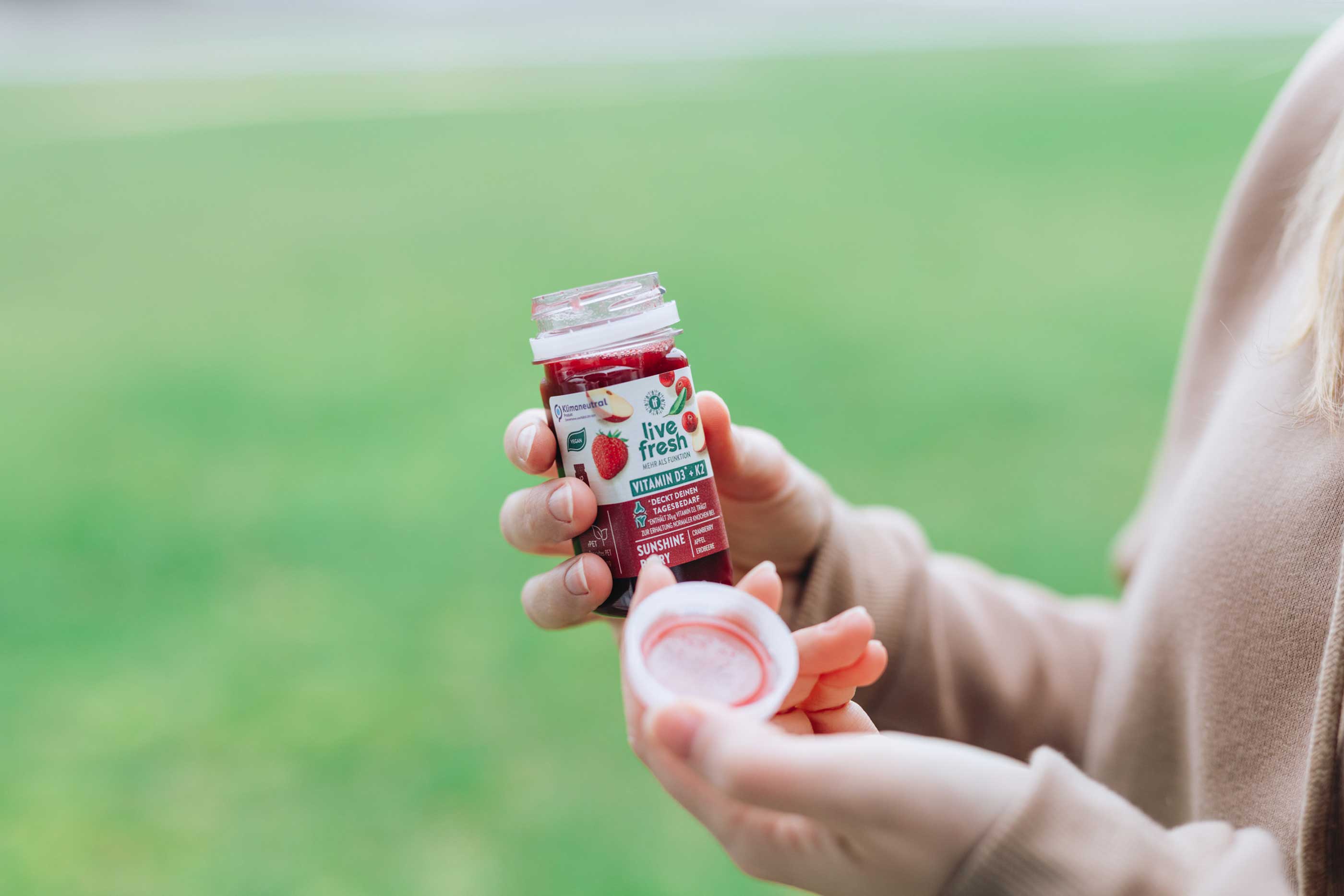 Person hält einen geöffneten Vitamin-D+K Shot namens Sunshine Berry von  LiveFresh in der Hand, im Freien vor grünem Hintergrund.
