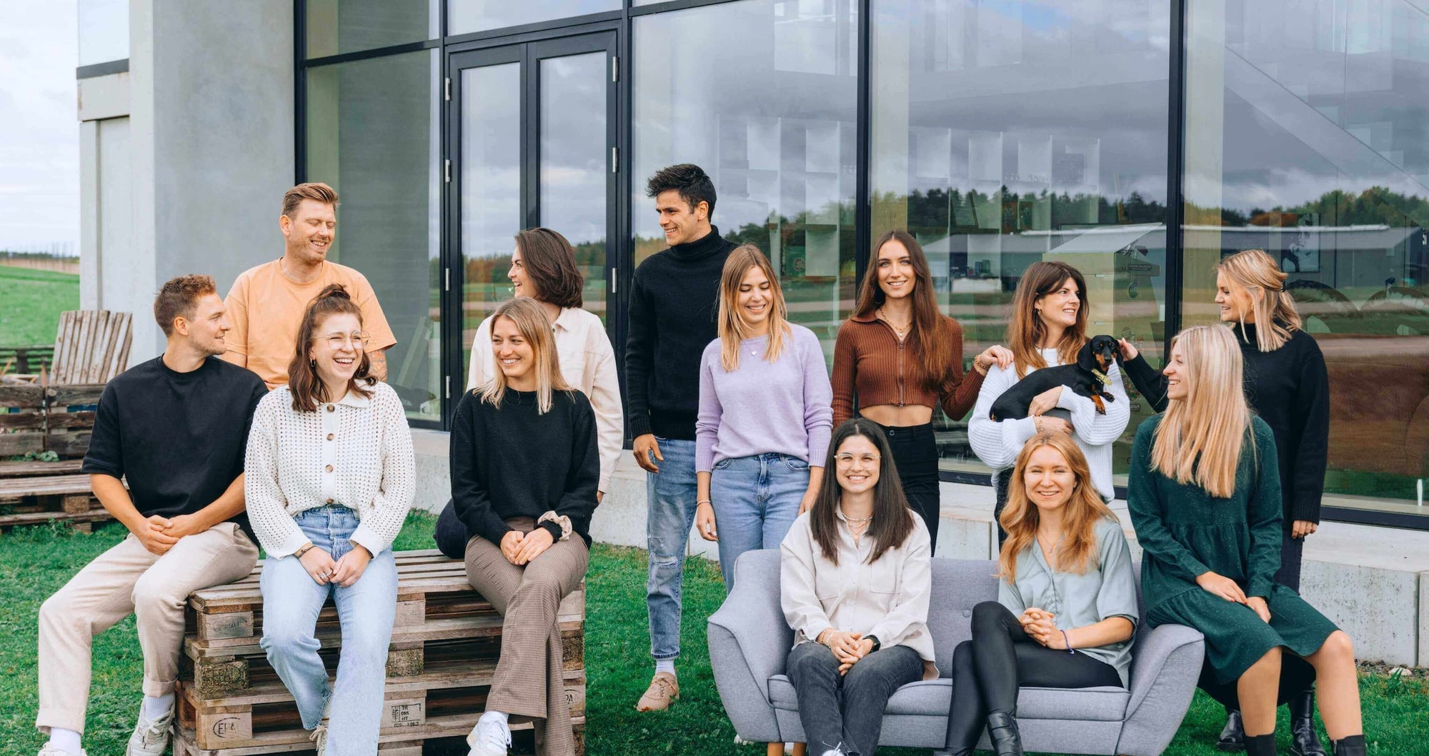Eine Gruppe von zwölf jungen Menschen sitzt und steht entspannt vor einem modernen Bürogebäude mit großen Glasfenstern. Sie lachen und unterhalten sich, während eine Person einen kleinen schwarzen Hund hält, der von einer anderen gestreichelt wird. Die Gruppe ist leger gekleidet, einige sitzen auf Paletten, andere auf einem grauen Sofa, mit einer grünen Wiese im Vordergrund und einem ländlichen Hintergrund.