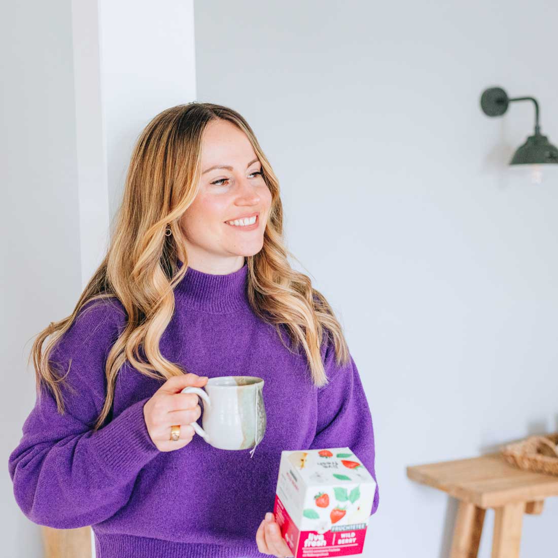 Frau in lila Pullover lächelt, während sie eine Tasse Tee trinkt und die LiveFresh Wild Berry Früchteteepackung in der Hand hält.