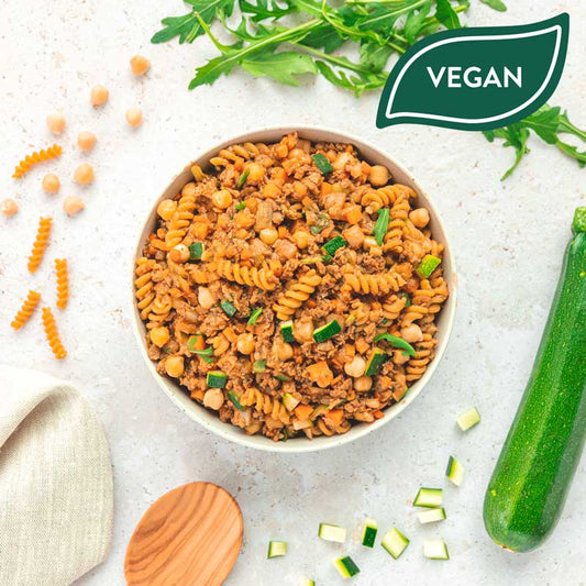 Bowl "Fusilli aus Kichererbsen – Toskanische Art" von LiveFresh mit Soja-Hack, Kichererbsen, Zucchini und Tomatensauce, serviert in einer weißen Schüssel. Drumherum liegen frische Zutaten wie Zucchiniwürfel, Kichererbsen, Fusilli-Nudeln und Rucola. Oben rechts ein grünes "Vegan"-Label.