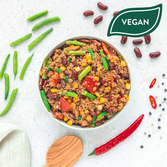 Eine Schüssel mit der "Gemüse Chili" Bowl von LiveFresh, bestehend aus Soja-Hack, roten Bohnen, Mais, grünen Bohnen und Tomaten, angerichtet auf einer hellen Oberfläche. Drumherum liegen frische Zutaten wie Chilischoten, grüne Bohnen, schwarze Pfefferkörner und ein Kochlöffel. Oben rechts ein grünes "Vegan"-Label.