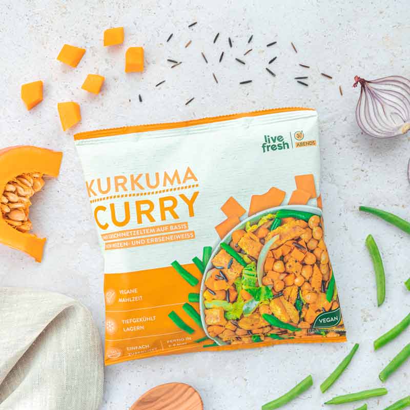 LiveFresh "Kurkuma Curry" Tiefkühlgericht in der orangenen Verpackung, umgeben von frischen Zutaten wie Kürbisscheibe und Kürbiswürfeln, grünen Bohnen, roter Zwiebel und Wildreis auf einer hellen Oberfläche. Ein Holzlöffel und ein Küchentuch liegen daneben.