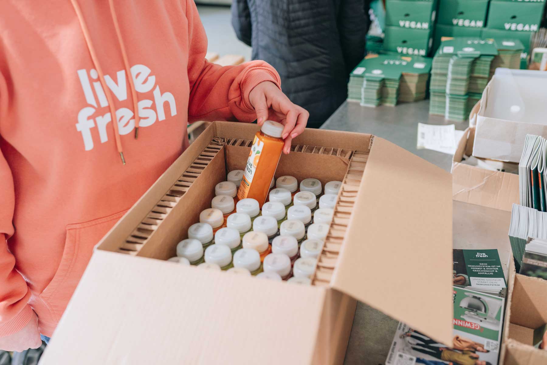 Zeigt eine Person in einem rosafarbenen Hoodie mit der Aufschrift „Live Fresh“, die eine Flasche Saft aus einem großen Karton entnimmt, der mit vielen weiteren Saftflaschen gefüllt ist. Im Hintergrund sieht man gestapelte Kartons mit der Aufschrift „Vegan“ sowie Broschüren und Verpackungsmaterial. Das Bild vermittelt eine Szene aus dem Versandprozess, wahrscheinlich der Vorbereitung von Saftbestellungen, und hebt die Organisation und die Sorgfalt bei der Lieferung der Produkte hervor.